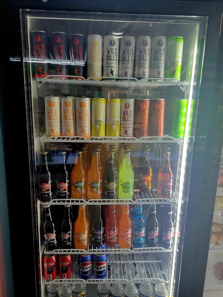 A glass-fronted refrigerated display cooler stocked with various colorful canned and bottled beverages organized on multiple shelves.