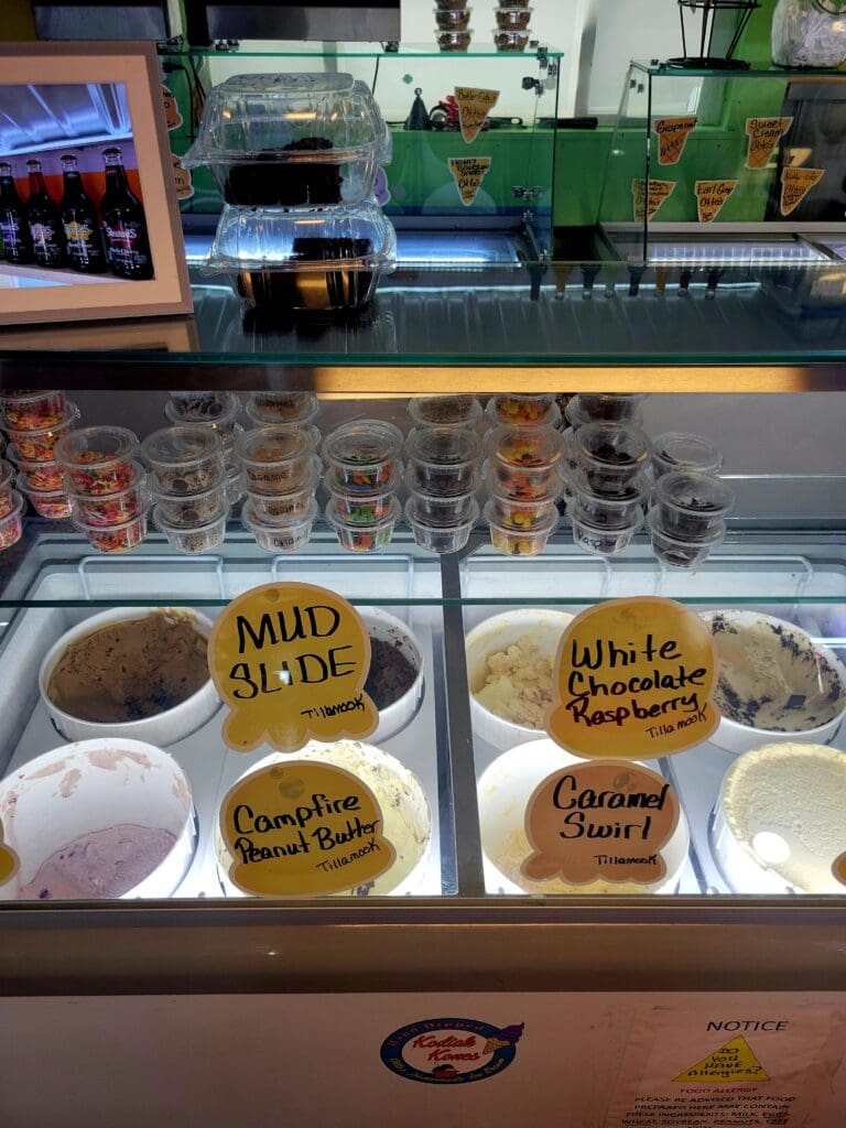 A refrigerated bakery display case showcases various baked goods and pastries with yellow price labels in what appears to be a commercial kitchen or bakery setting.