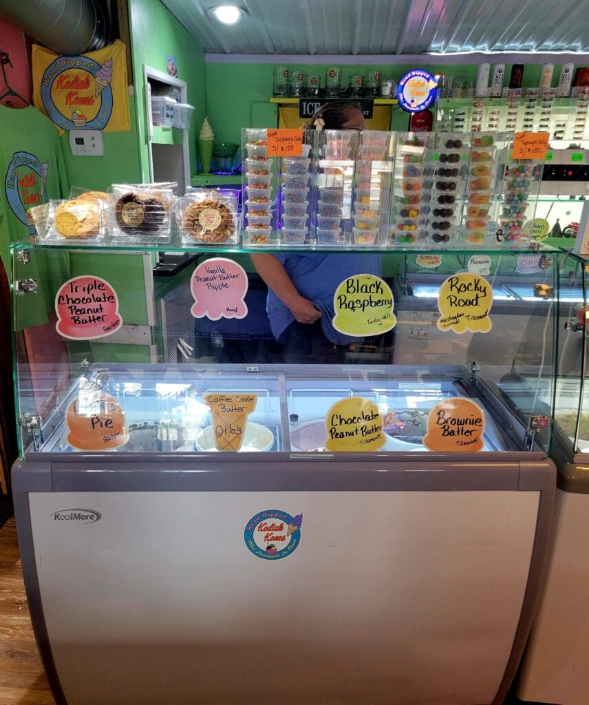 A refrigerated ice cream display case in a bright green candy or ice cream shop with various flavors visible and a staff member behind the counter.