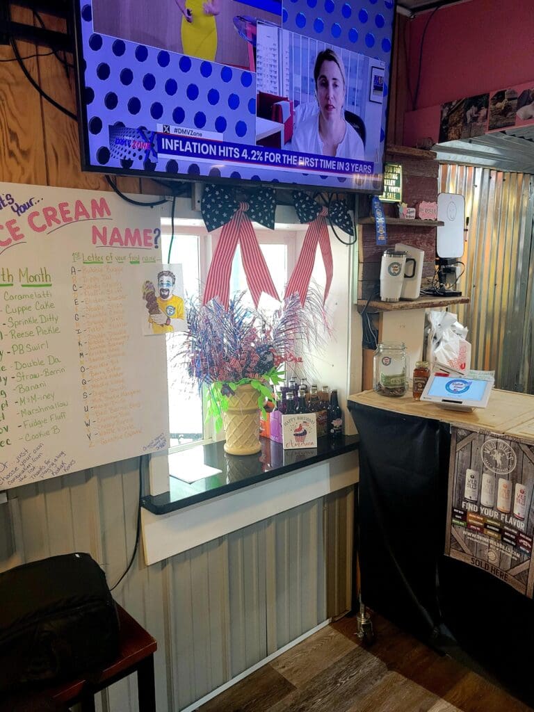 A small workshop or office space with purple-blue lighting, a wall-mounted television display, a windowed display case with plants and decorations, and various workspace elements including a whiteboard and desk area.