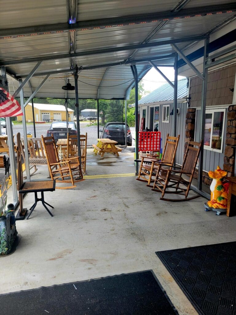 An industrial workshop or storage facility with wooden rocking chairs, tools, and equipment arranged throughout, lit by natural light and fluorescent overhead lighting.