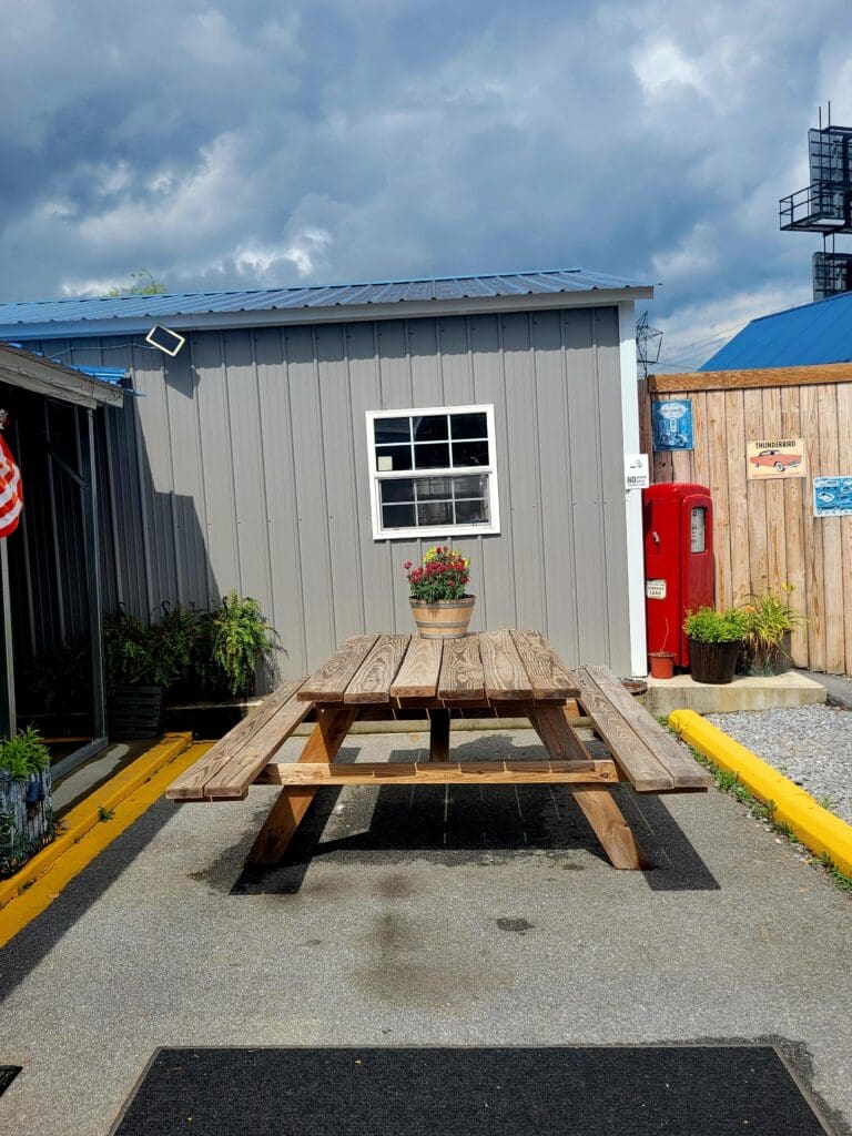 A wooden picnic table sits on a painted parking space in front of a small industrial workshop or storage building with blue and beige siding under a partly cloudy sky.