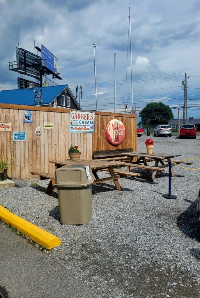 A carnival or fairground setup showing a blue wooden equipment trailer and mechanical ride components parked in a gravel lot under partly cloudy skies.