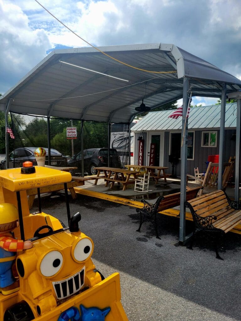 A backyard play area with yellow playground equipment, picnic tables, and a covered canopy structure on a sunny day.