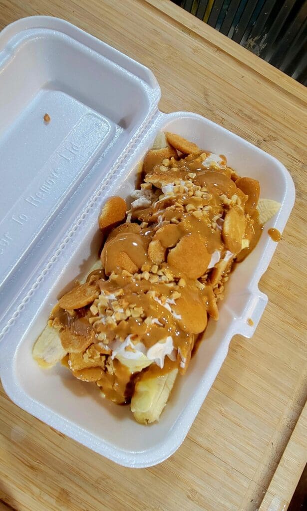 A close-up overhead view of loaded French fries topped with melted cheese and seasonings served in a white disposable container.