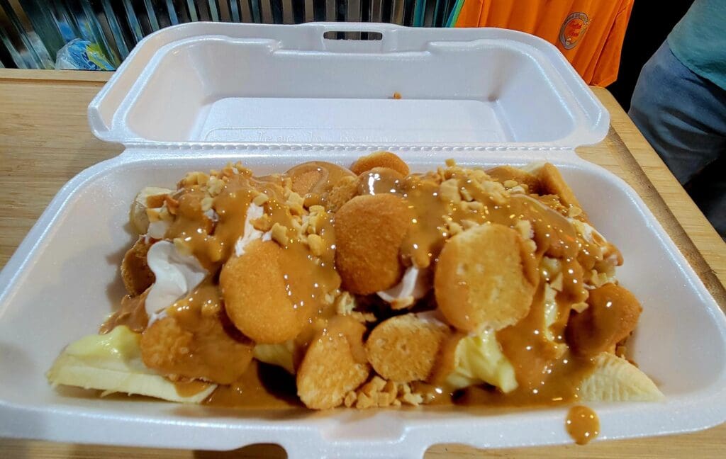 A close-up of fried food served in a white disposable container on a wooden surface, likely at a casual dining or carnival venue.