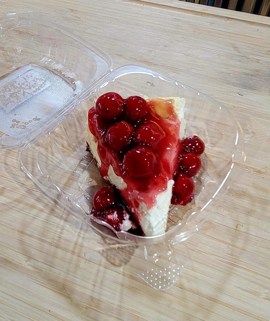 A slice of strawberry cheesecake with fresh red berries sits in a clear plastic container on a wooden table.