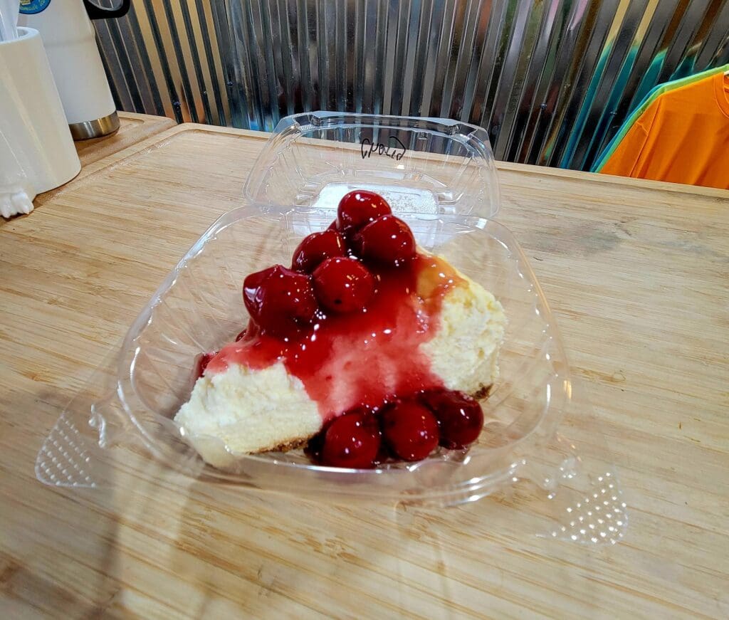 A fresh dessert topped with whipped cream and bright red currants served in a clear plastic container on a wooden table.