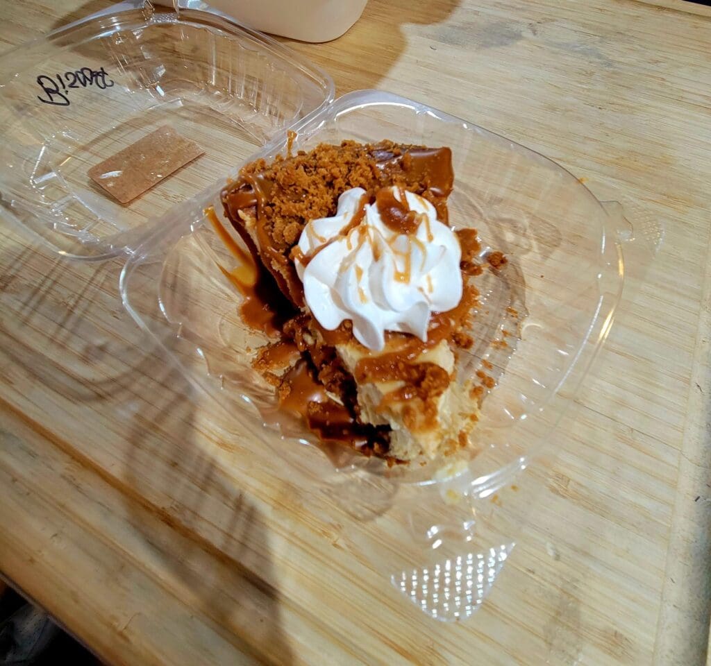 A close-up of a golden-brown apple pie served in a clear plastic container topped with whipped cream on a wooden table.