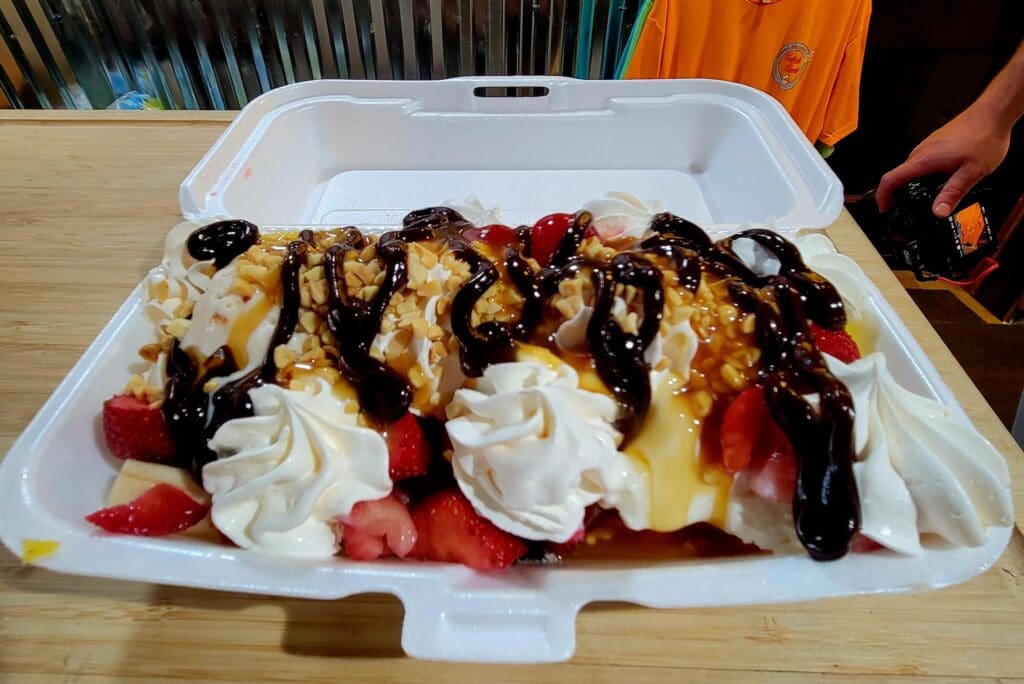 An overhead view of an elaborately plated dessert with whipped cream, fresh berries, chocolate drizzle, and pastry garnishes served in a white container.