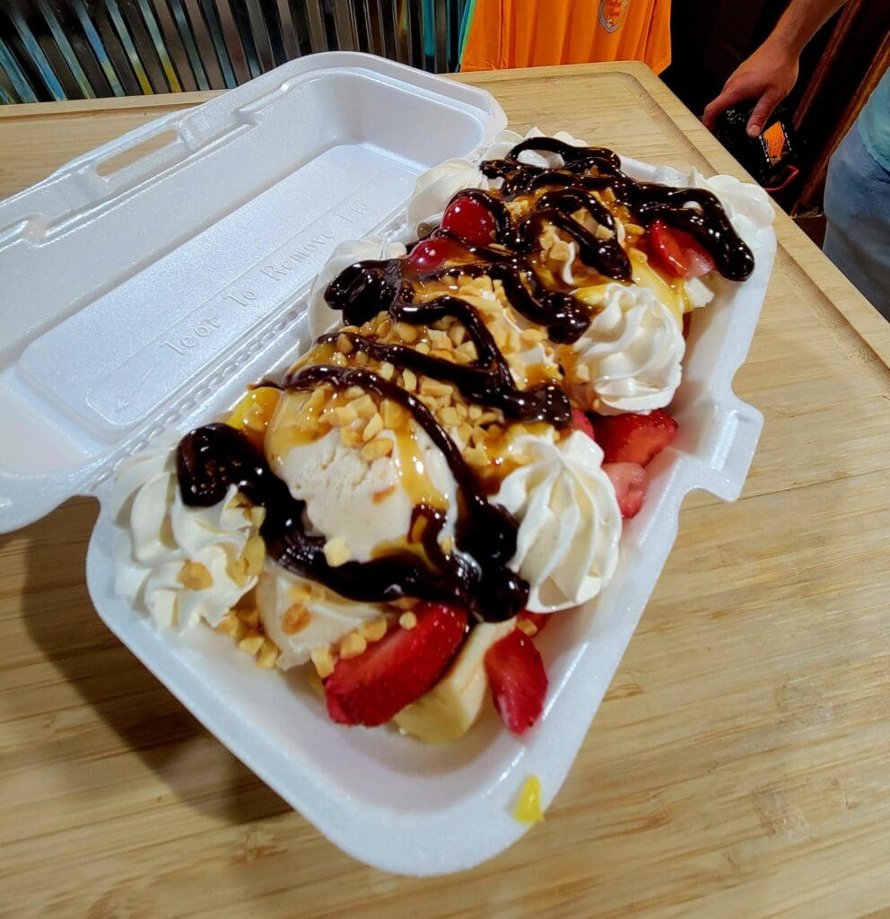 An overhead view of a decadent dessert platter featuring whipped cream, berries, chocolate drizzle, and waffles served in a white disposable container.