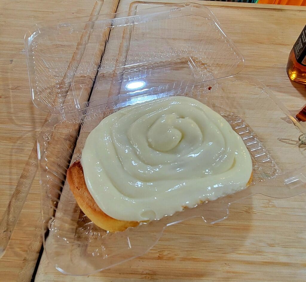 A freshly baked cinnamon roll with white frosting sits on a wooden surface, partially wrapped in clear plastic.