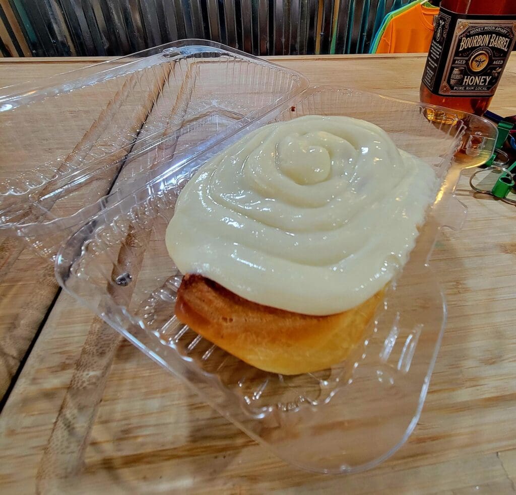 A freshly baked cinnamon roll with thick white frosting sits in a clear plastic bakery container on a wooden table.