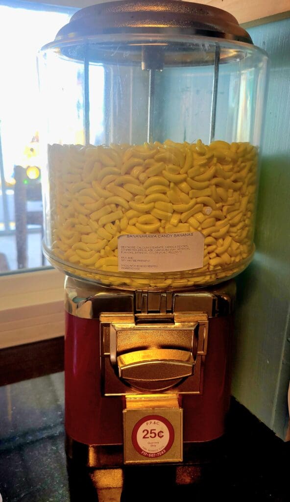 A vintage-style red and wooden gumball machine filled with yellow gumballs, photographed indoors against a light blue wall.