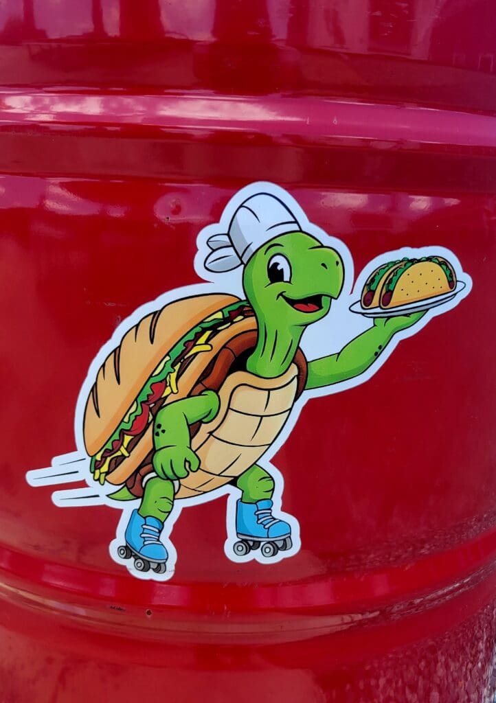 A cheerful cartoon turtle character wearing a chef's hat and holding a taco is depicted as a sticker on a bright red vehicle surface.