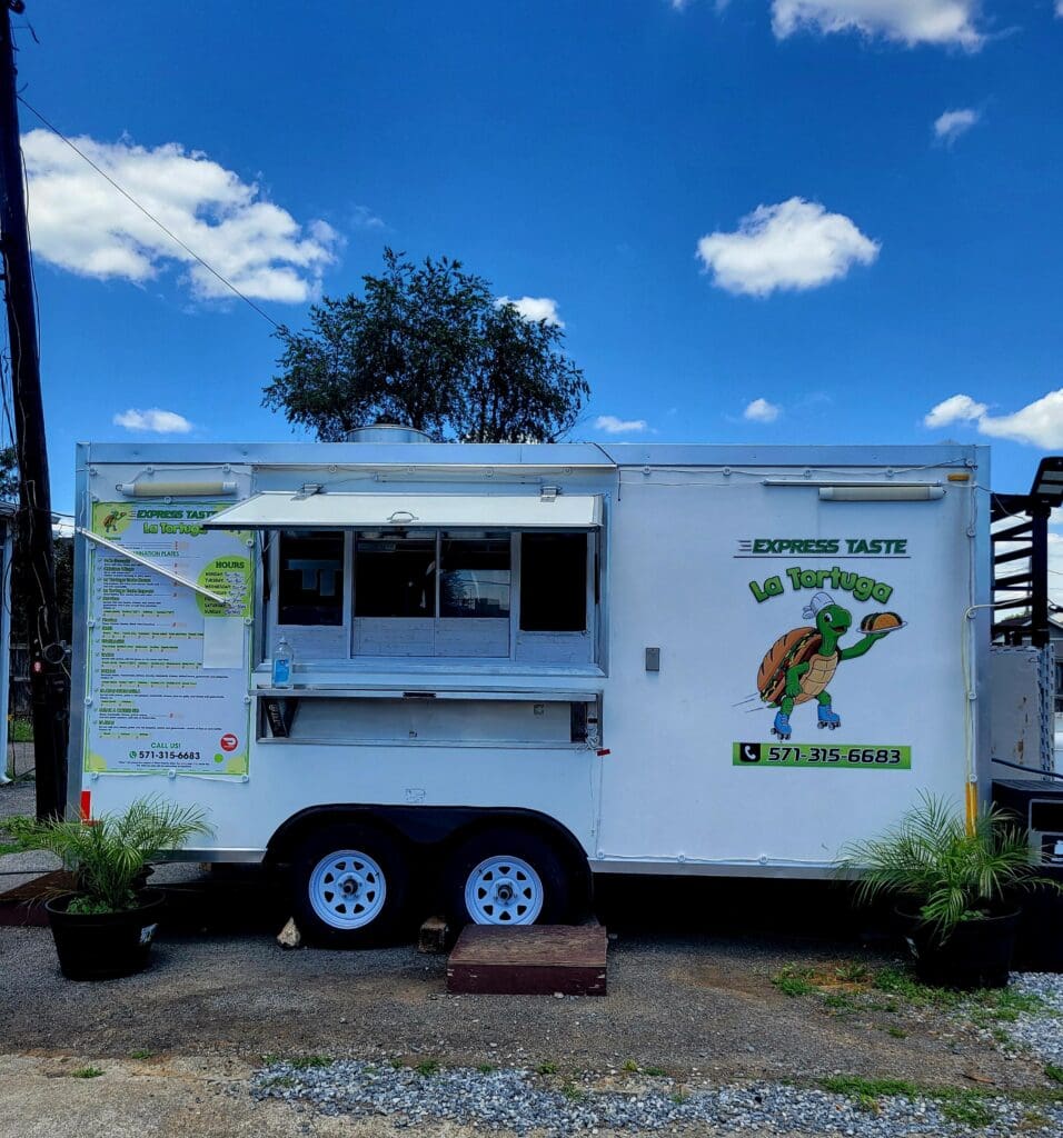 A white food service trailer with colorful graphics sits parked on gravel under a bright blue sky with scattered clouds.