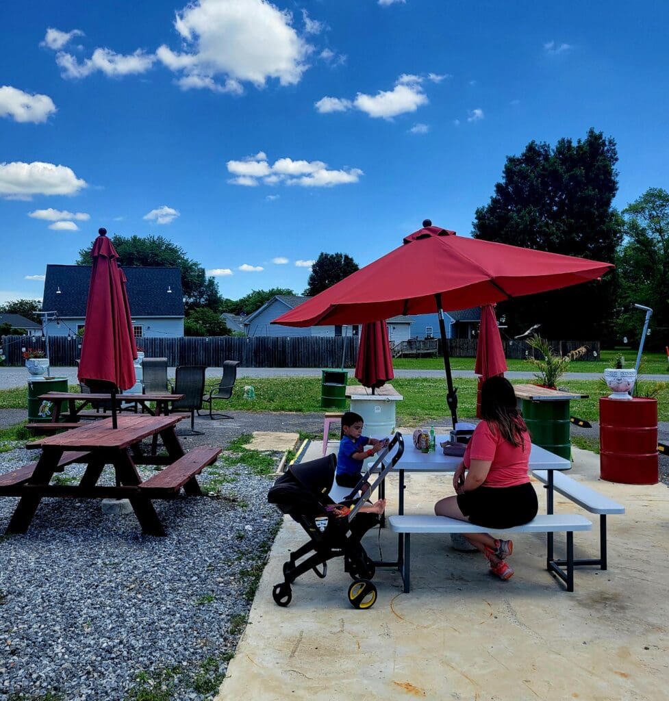 A peaceful community park or picnic area features red canopies and picnic tables where a few people gather on a sunny day with clear blue skies.