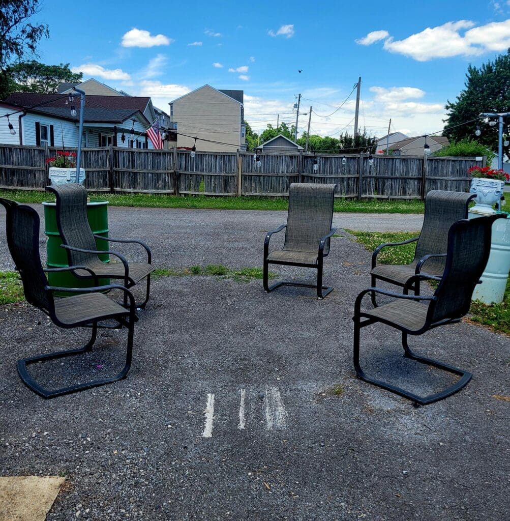 A peaceful outdoor community seating area with four black cantilever chairs arranged in a circle on an asphalt pad in a residential neighborhood on a sunny day.