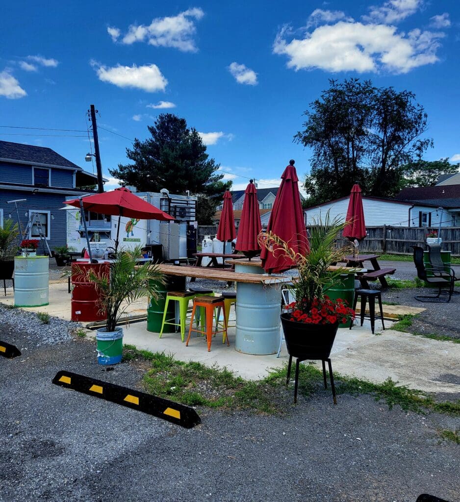 A sunny outdoor market or community gathering space with red umbrellas, picnic tables, and residential homes in the background under clear blue skies.