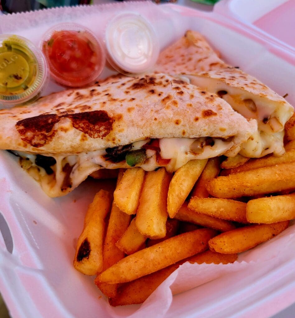 A wrapped sandwich filled with vegetables and cheese is served on a pink plate alongside golden french fries and small condiment containers.