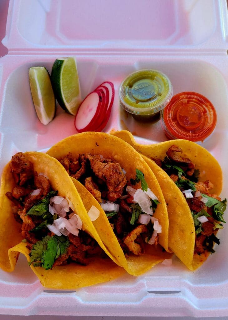 Three golden-fried tacos filled with meat and fresh toppings are arranged in a white disposable container with colorful condiments and garnishes on the side.
