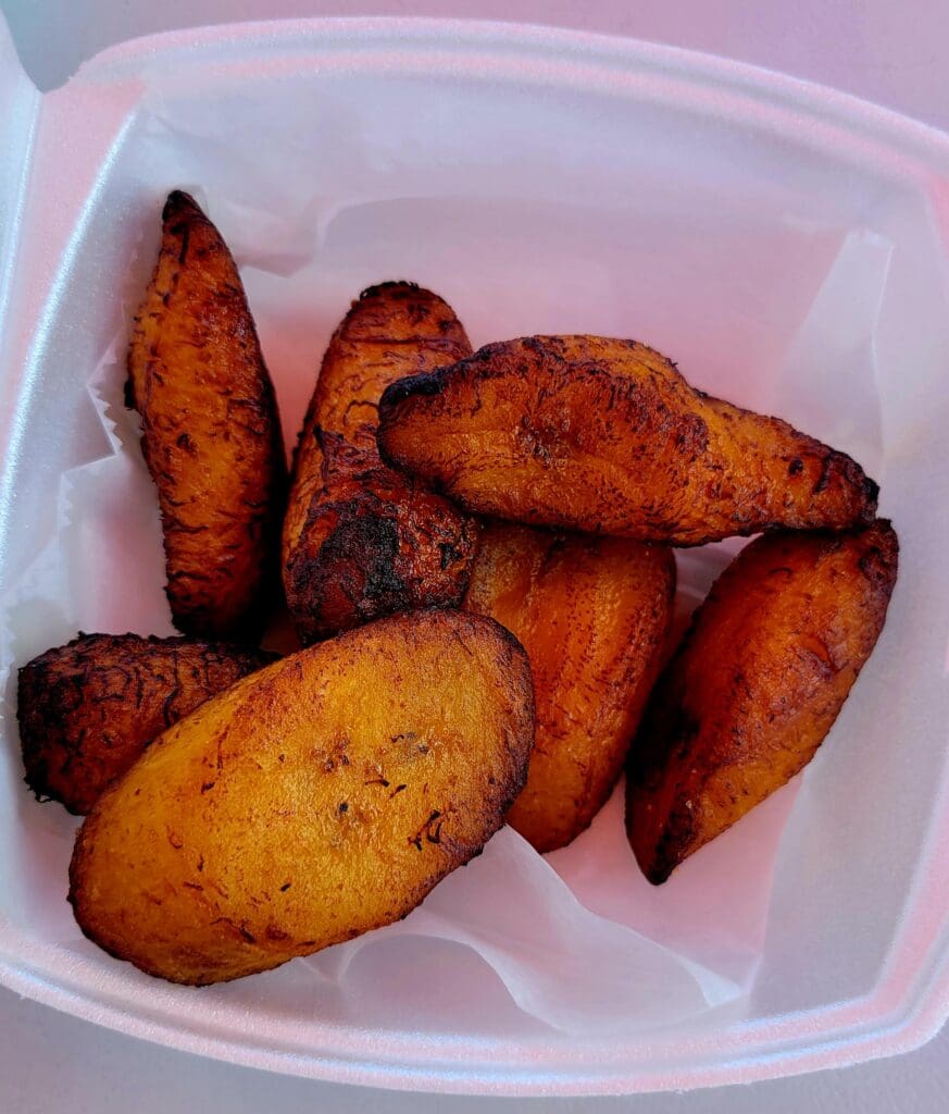 A close-up photograph of golden-brown fried plantains arranged in a white plastic takeout container.