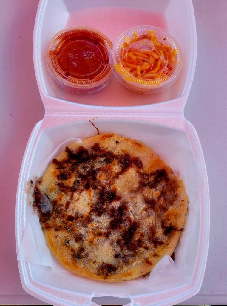 A personal pizza with melted cheese and toppings is displayed in a pink takeout container alongside two cups of red dipping sauce.