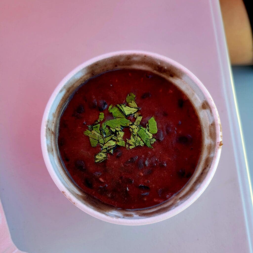 A vibrant deep red beet soup garnished with fresh green herbs served in a white bowl on a pink surface.
