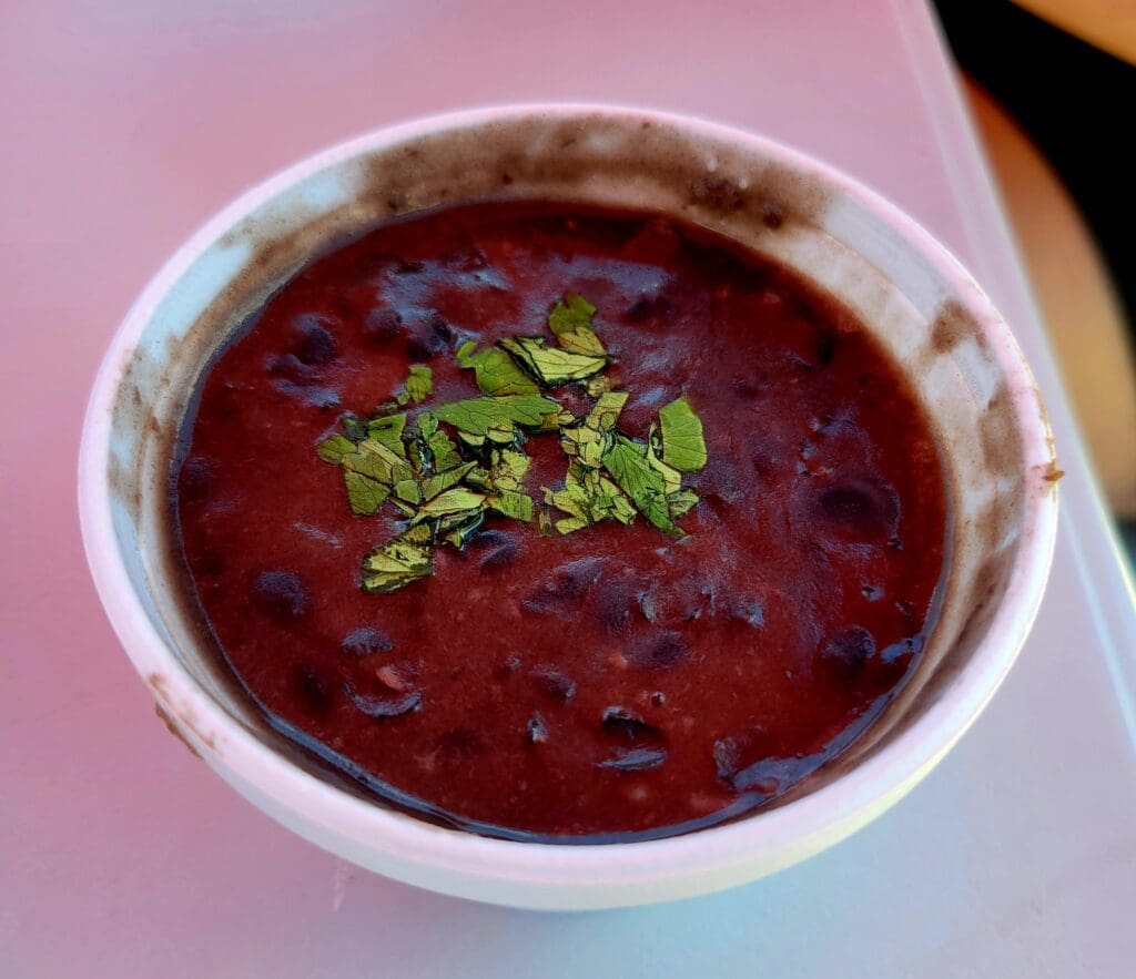 A vibrant deep red beet soup served in a white bowl, garnished with fresh green herbs on a pink table surface.