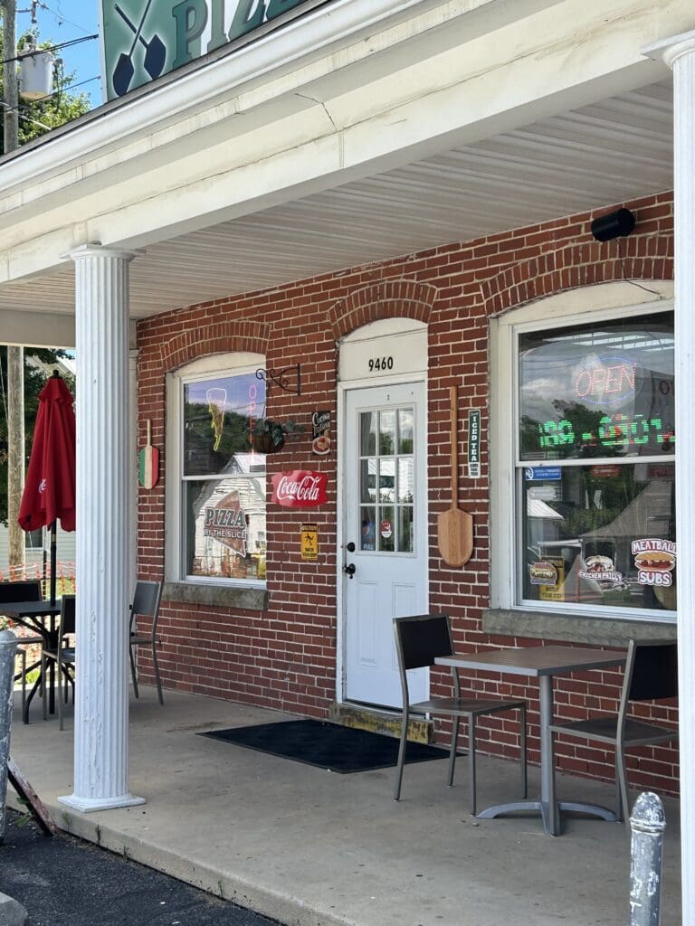 A charming brick storefront with white columns features a vintage entrance, display windows, and vintage furniture suggesting a small-town shop or antique store.