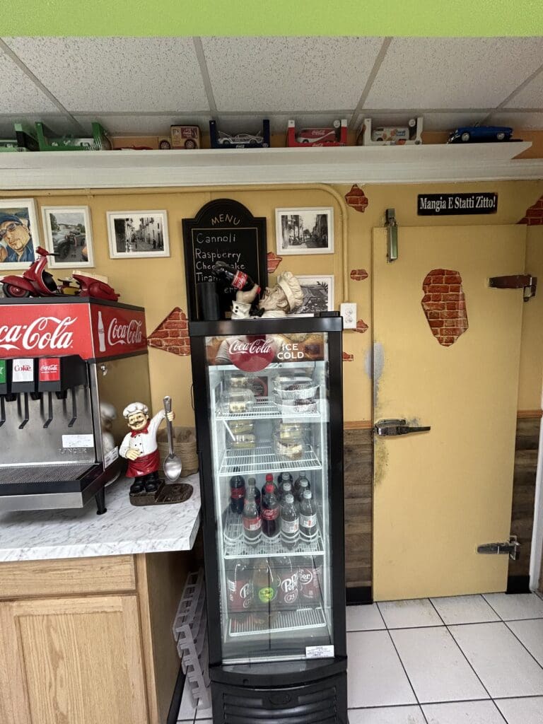 A retro-style small cafe or diner interior with yellow walls, vintage Coca-Cola signage, a beverage cooler, and classic soda fountain counter.