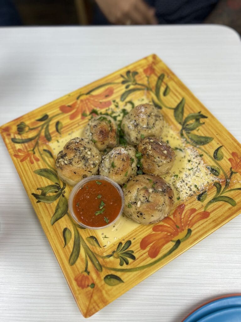 A decorative yellow plate with herb patterns holds four cooked meatballs with a small bowl of red sauce in the center.