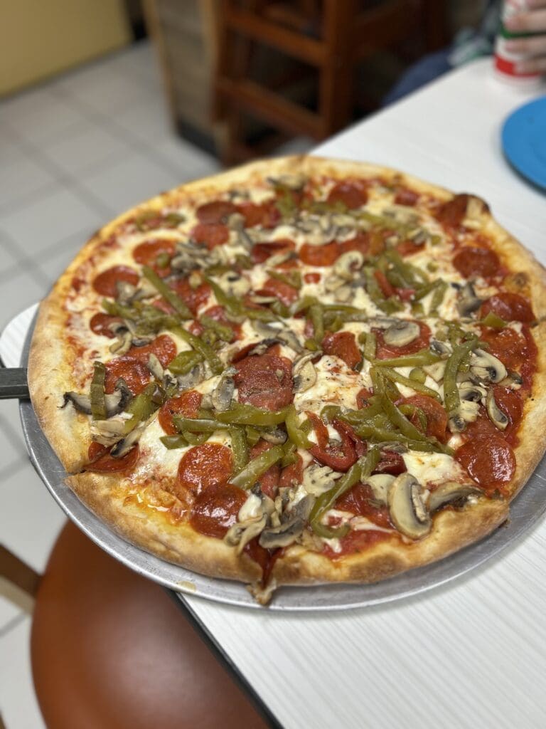 A freshly served pepperoni and vegetable pizza on a white plate with a person's hand visible at the bottom of the frame in an indoor setting.