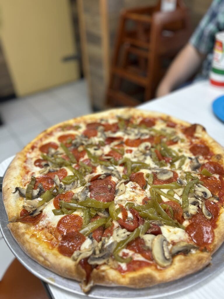 A freshly prepared pepperoni pizza with mushrooms and green peppers sits on a plate in an indoor kitchen setting.