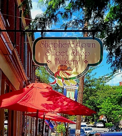 A charming sweet shop storefront with a vintage-style sign and bright red umbrella on a sunny street lined with trees.