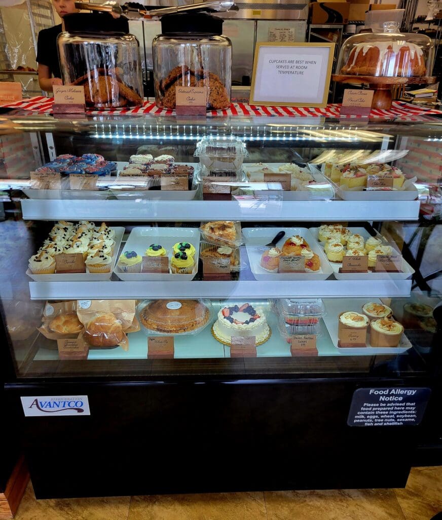 A glass-fronted bakery display case showcases an assortment of baked goods, pastries, and desserts arranged on multiple shelves.