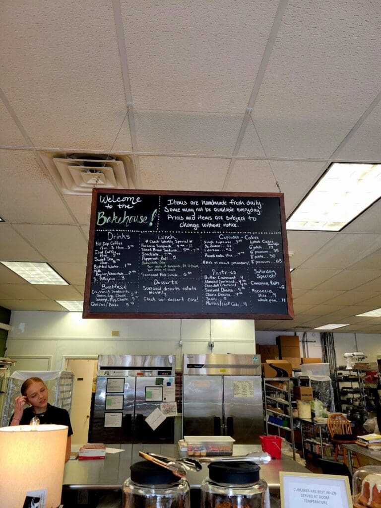An indoor workshop or classroom space with a large chalkboard menu mounted on the ceiling, fluorescent lighting, and various storage and furniture elements visible.