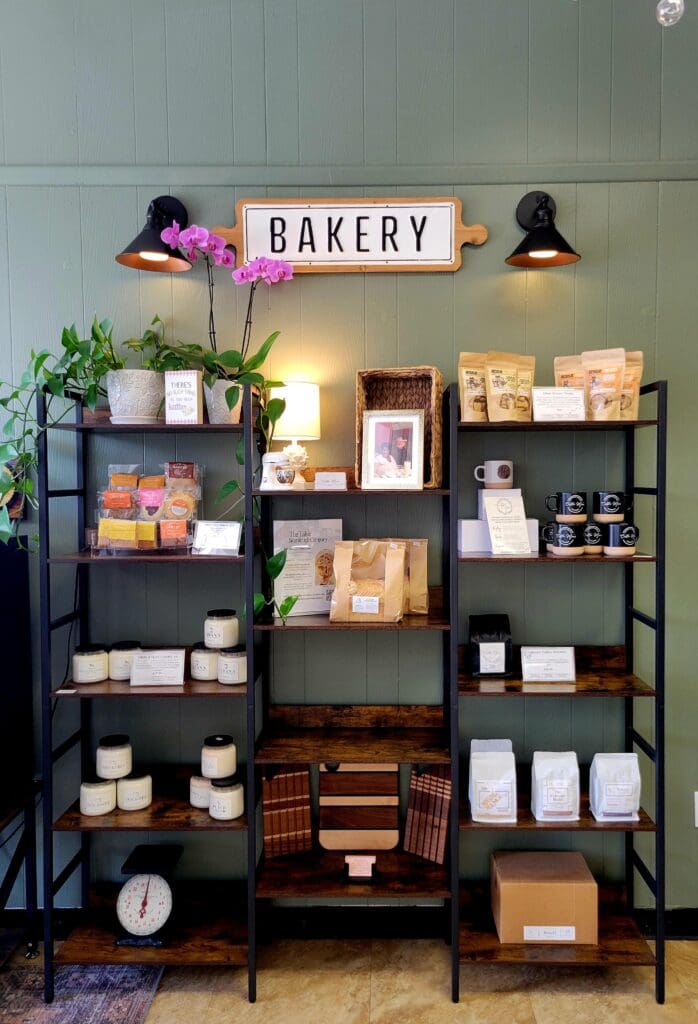 A well-organized bakery retail display with black metal shelving units stocked with packaged goods, plants, and products, illuminated by a glowing pink neon 'BAKERY' sign above.