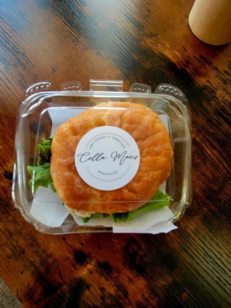 A freshly baked pastry with a golden-brown exterior sits in a clear plastic bakery box with a white circular label, displayed on a warm wooden surface.