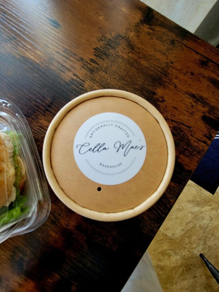 A round beige food container with a white label sits on a wooden table next to packaged food items.