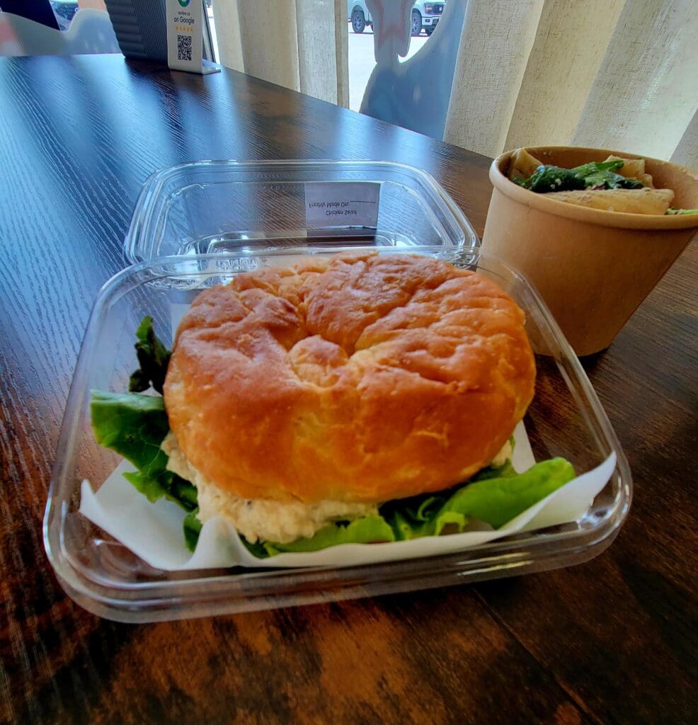 A fresh croissant sandwich with green lettuce on a wooden table in what appears to be a casual dining or takeout setting.
