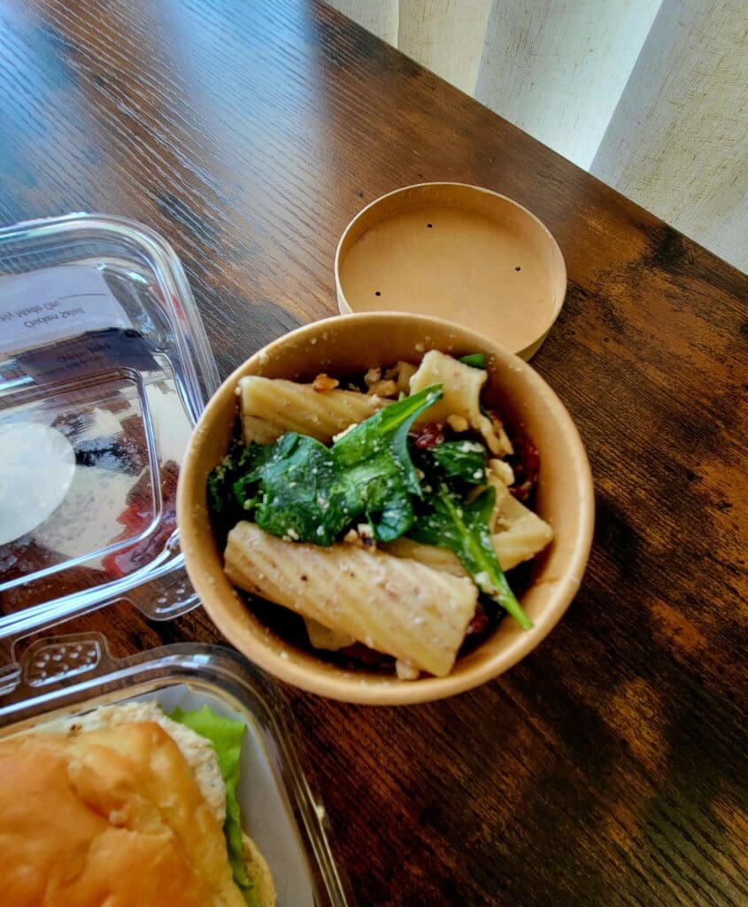 An overhead view of an Asian noodle dish served in a wooden bowl with fresh greens, accompanied by a beverage glass and takeout containers on a wooden table.