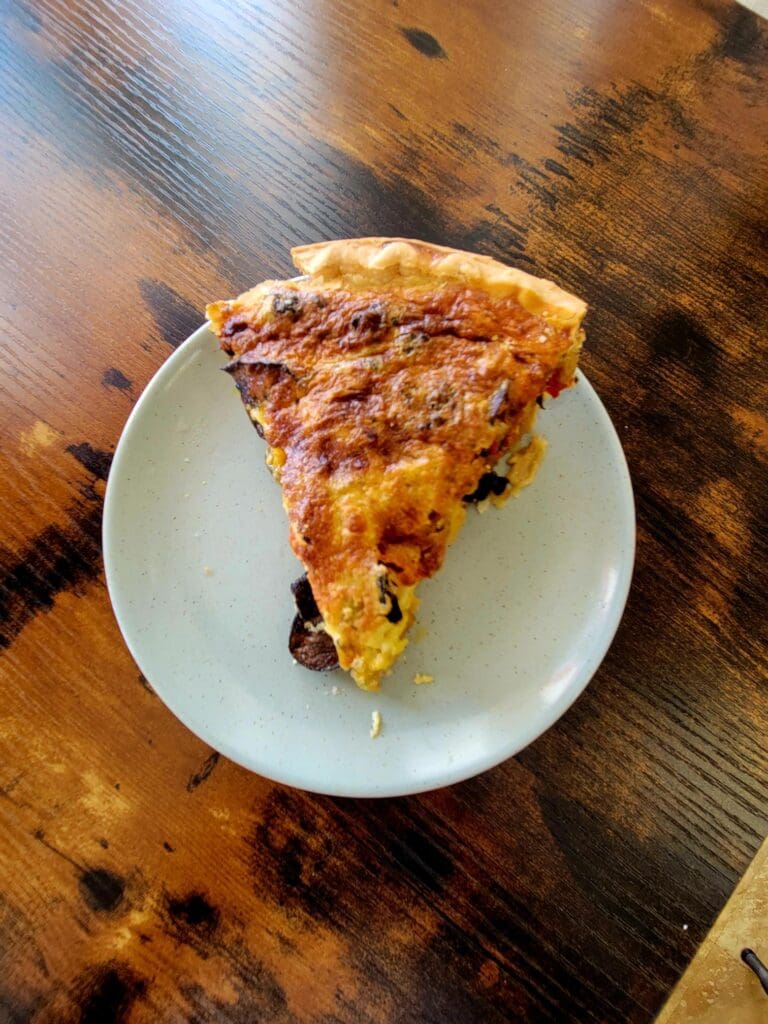 A slice of pizza with melted cheese and toppings sits on a white plate atop a rustic wooden table.