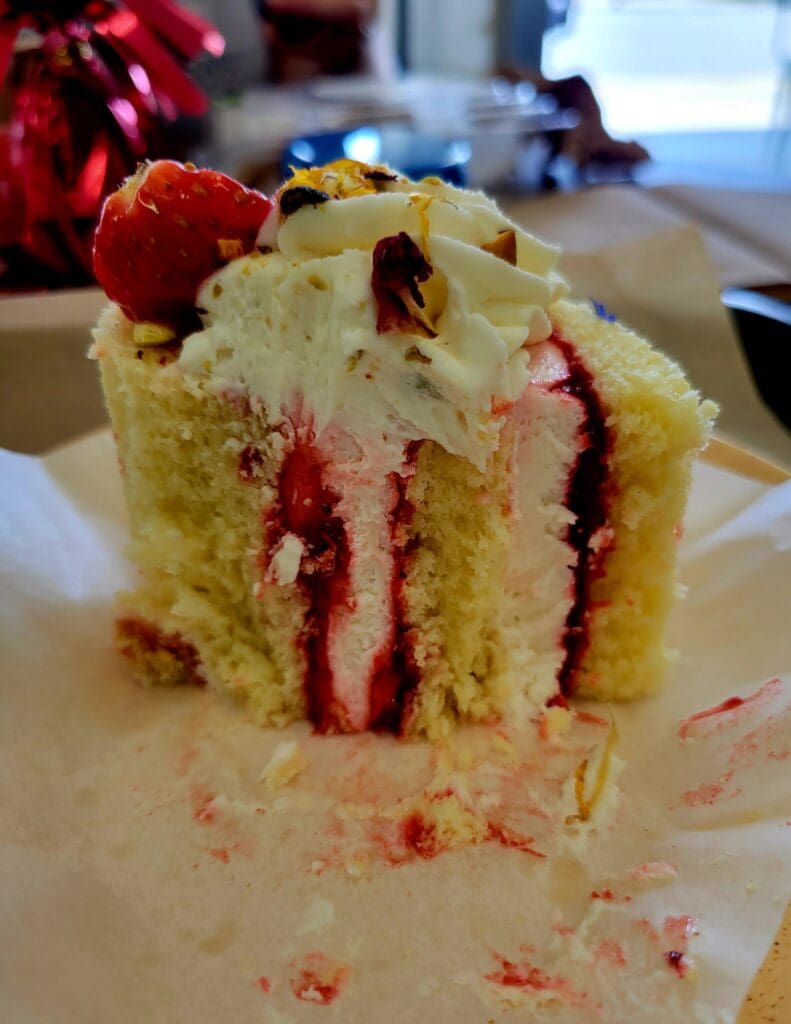 A close-up photograph of a slice of multi-layered vanilla cake with strawberry filling and fresh strawberry garnish on a white plate.