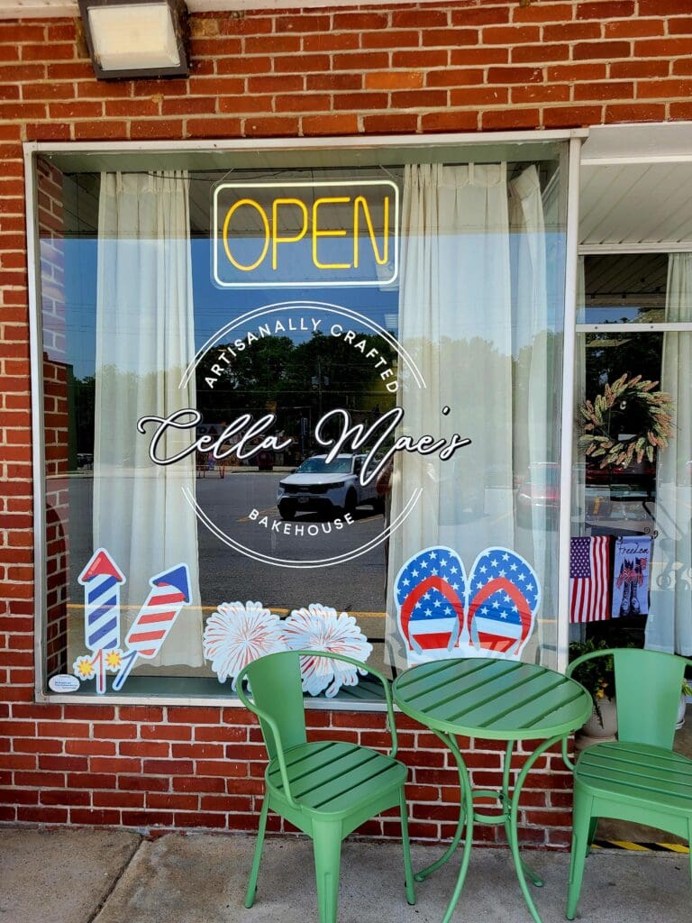 A charming brick storefront with an 'OPEN' neon sign, green chairs, and holiday wreath decoration in the window display.