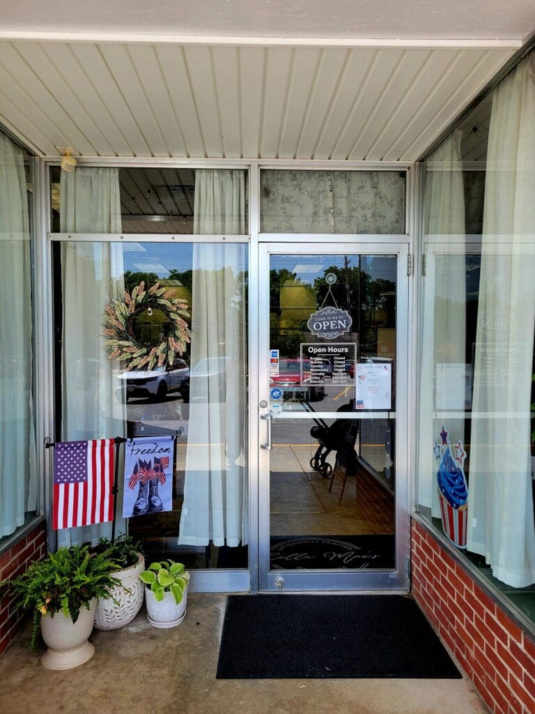 The entrance to a small business storefront featuring glass doors, an American flag, and welcoming potted plants on a brick facade during daytime.