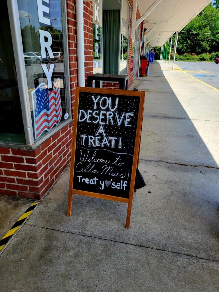 A wooden A-frame sandwich board sign on a brick storefront sidewalk advertises treats and specials during daytime.