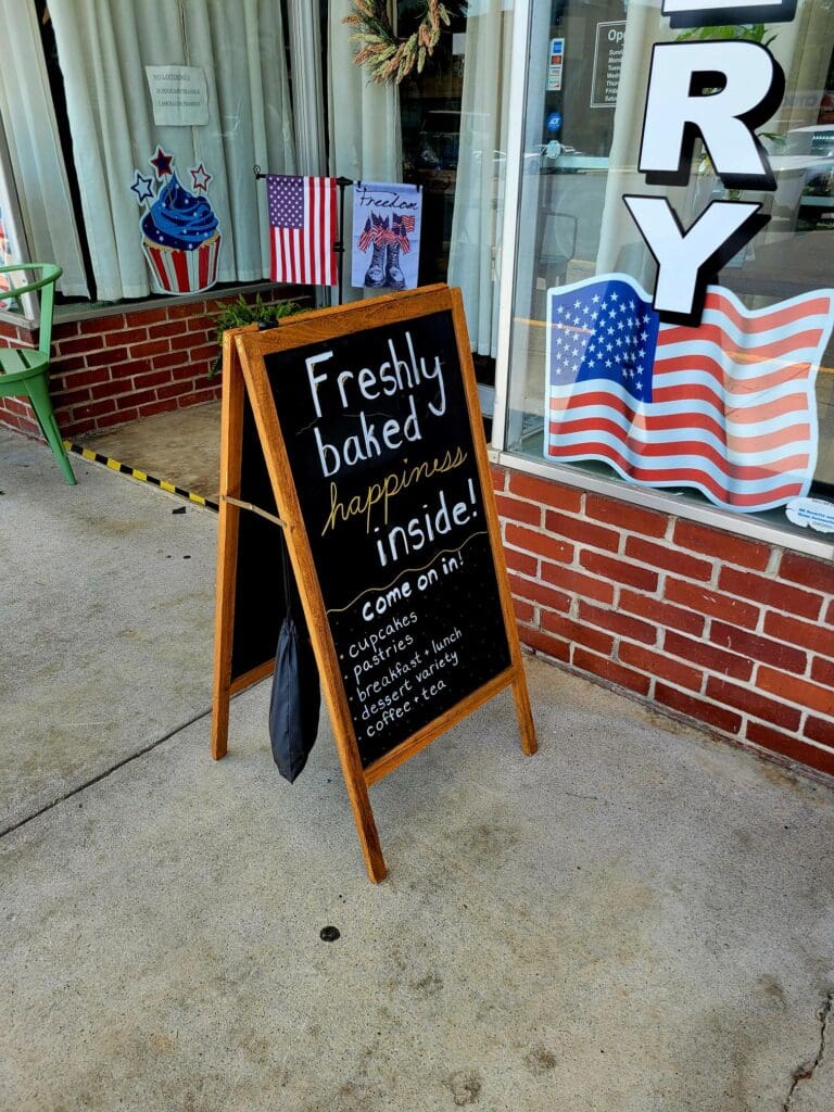 A storefront displays a sandwich board advertising fresh baked goods and fried items, decorated with American flags on a brick building facade.