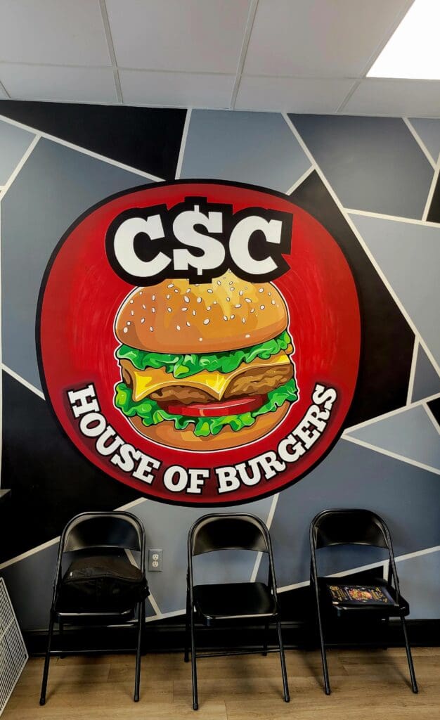 A large red circular burger restaurant sign for 'CSC House of Burgers' hangs from the ceiling above black dining chairs in a modern indoor venue.