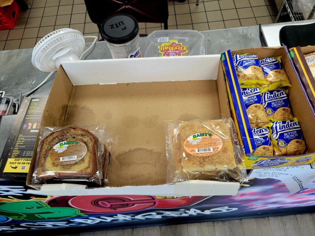An overhead view of a cardboard box containing various packaged food items including baked goods, cereal boxes, and disposable plates on a kitchen counter.